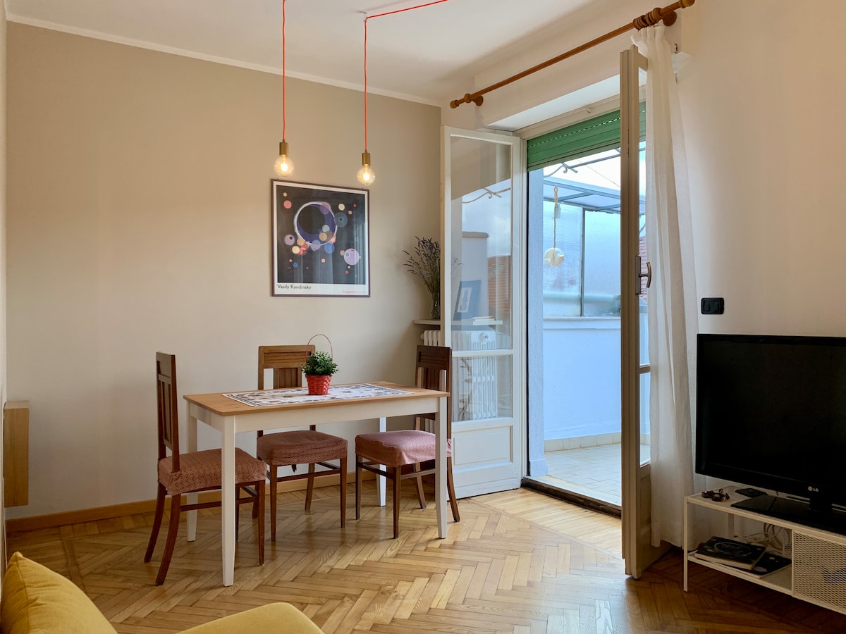 A dining area features a white table surrounded by four wooden chairs, with a small potted plant at the center. A sliding glass door leads to a balcony, bringing in natural light. A television is positioned nearby, enhancing the functional living space.