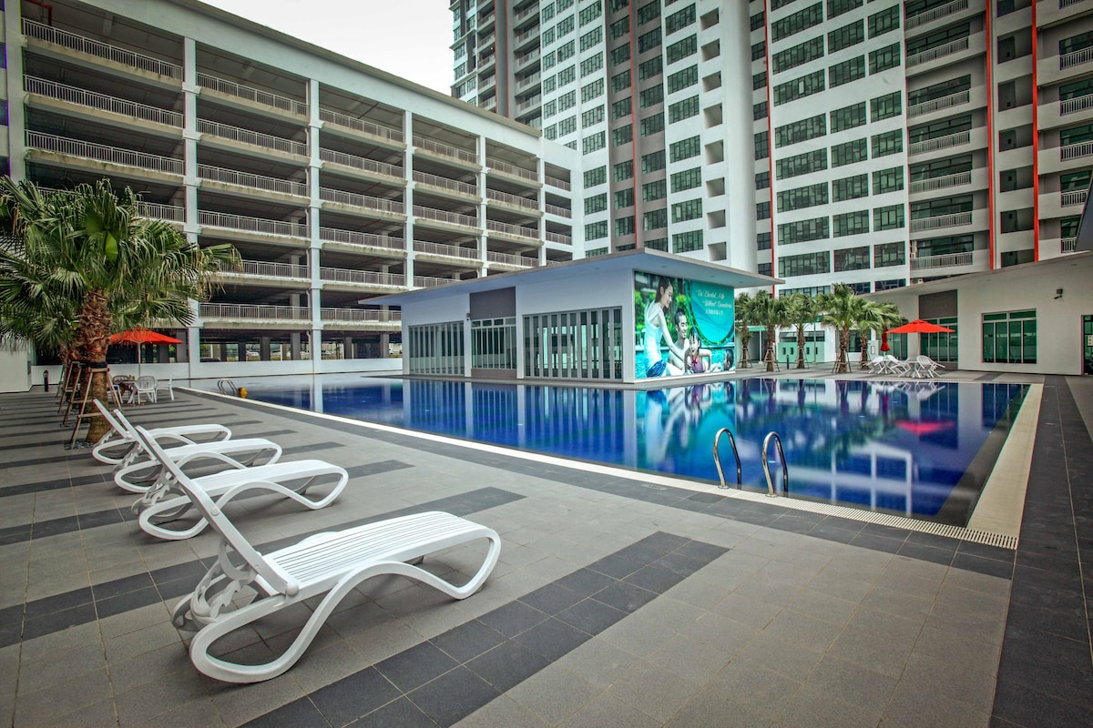 A large swimming pool is surrounded by modern apartment buildings, featuring lounge chairs positioned around the deck. Colorful umbrellas provide shade near the water's edge, and the pool's surface reflects the surrounding architecture.