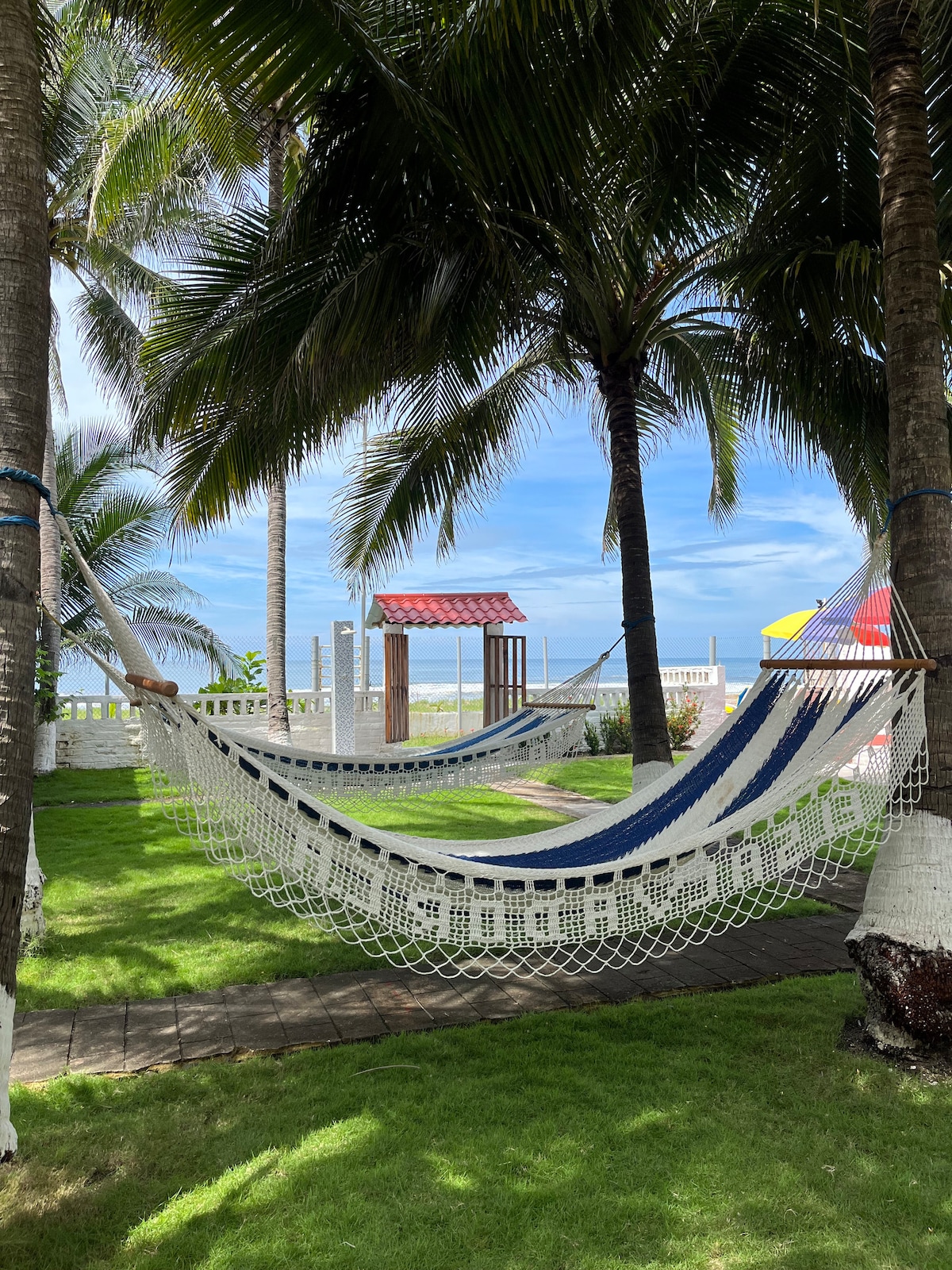 A hammock is suspended between palm trees in a grassy area, creating a relaxing space. A gazebo can be seen in the background, with a clear view of the sky and distant shoreline. The vibrant green grass enhances the tranquil setting.