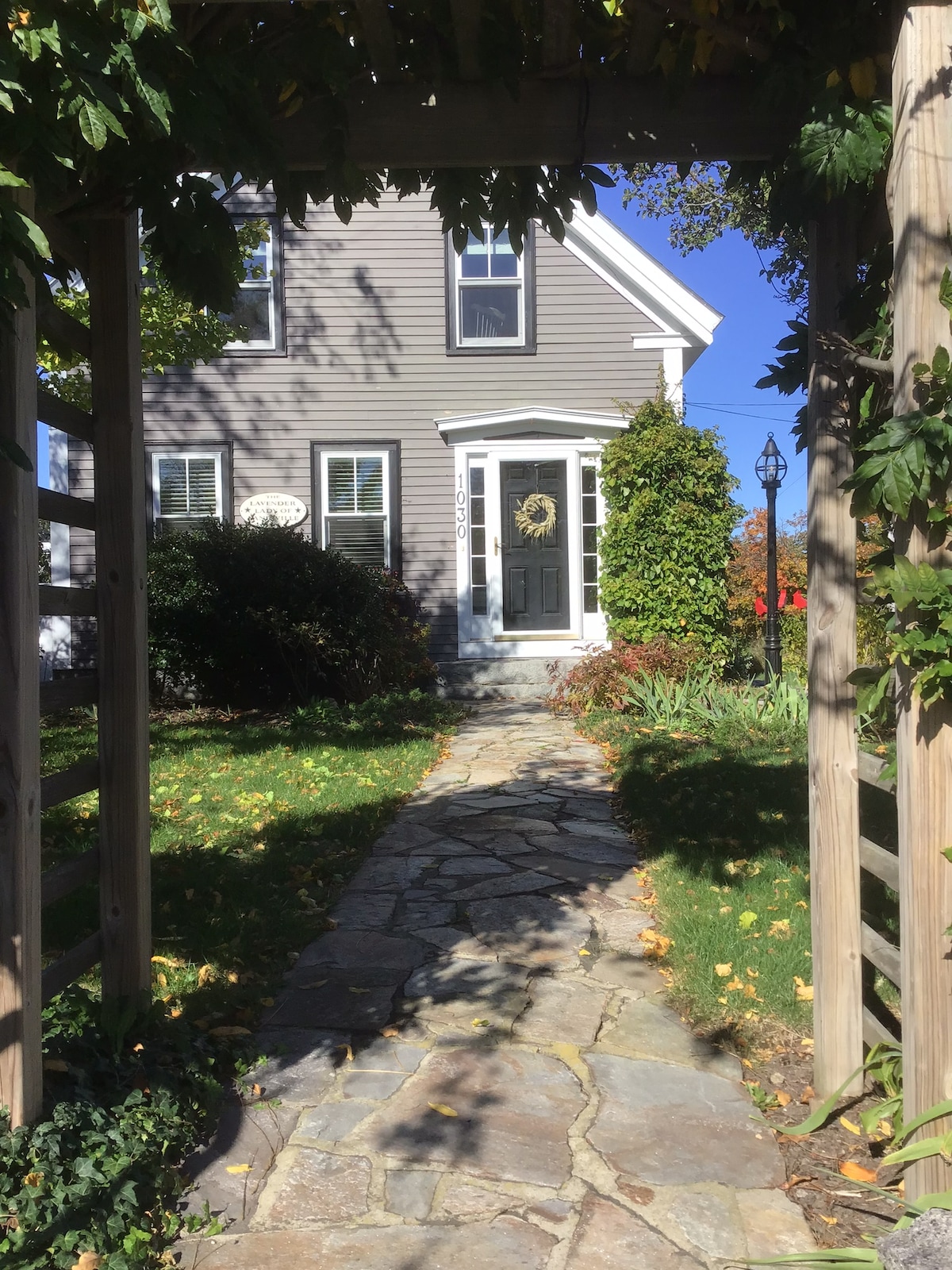 A stone pathway leads to a charming gray house framed by lush greenery. The entrance is enhanced by a decorative wreath on the door, and bright sunlight illuminates the inviting front yard. Clear blue skies complement the home’s welcoming facade.