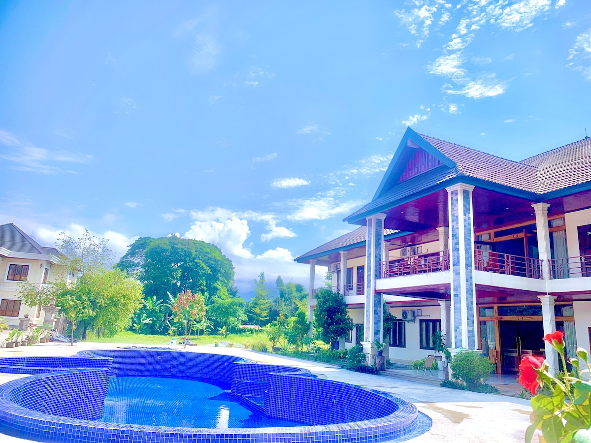 A spacious outdoor area features a blue tile swimming pool surrounded by lush greenery. The two-story building showcases a traditional architectural style with large windows and a covered balcony. The sky above is bright with scattered clouds.