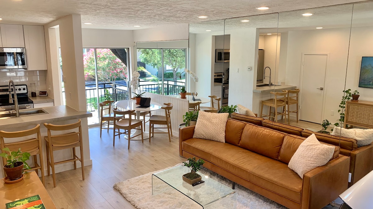 An open living area is showcased, featuring a spacious tan sofa with decorative pillows and a glass coffee table. A dining table with wooden chairs is nearby, and large windows allow natural light to fill the room, offering views of the surrounding greenery.