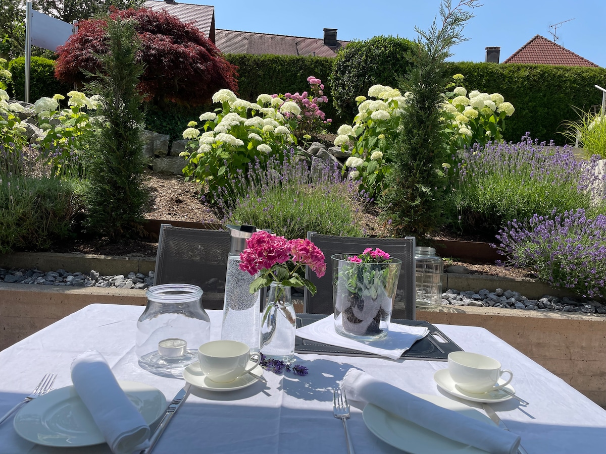 A dining table is set under sunlight, featuring white tablecloths and two teacups. Fresh flowers in vases are displayed, with vibrant greenery and blooming hydrangeas and lavender in the background, creating an inviting outdoor dining space.