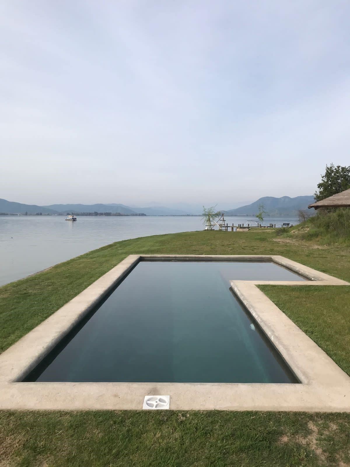 A private swimming pool is situated alongside a lake, featuring a sleek design with clean lines. Lush green grass surrounds the pool, complemented by distant mountains and a calm water surface. The serene landscape invites relaxation and enjoyment of nature.