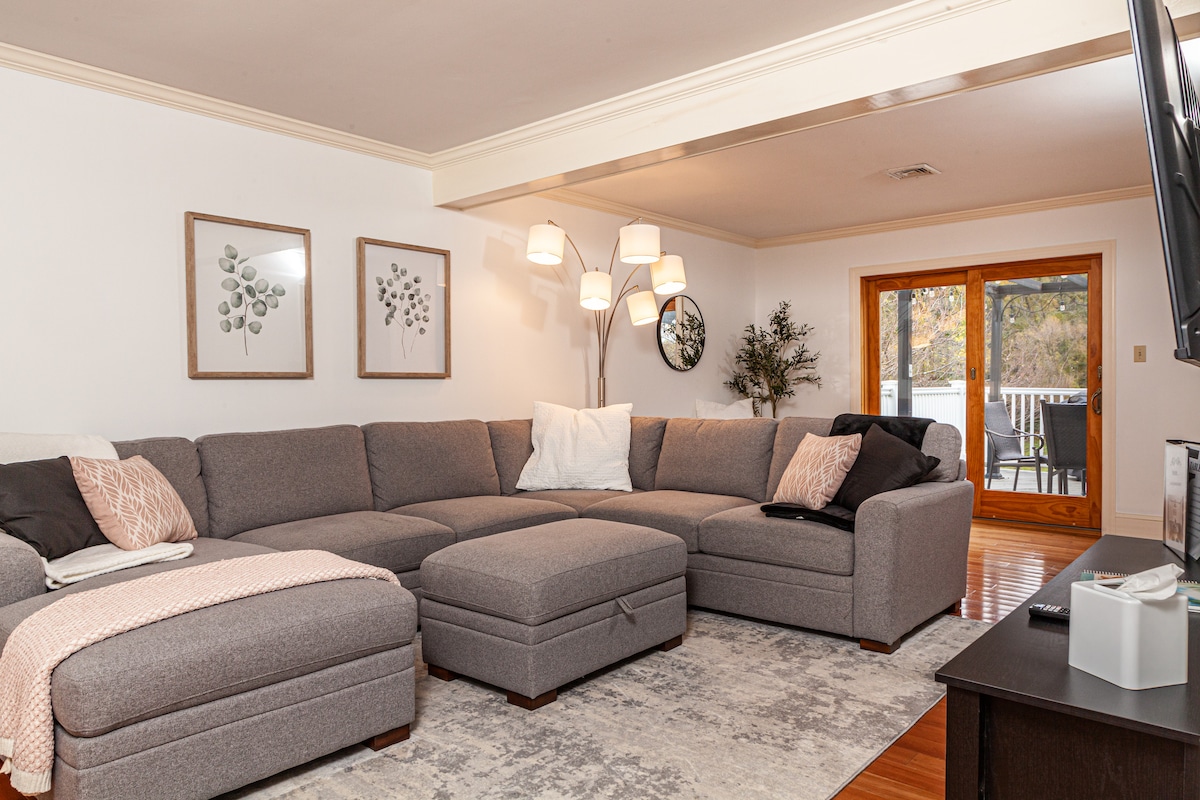 A spacious living room is presented, featuring a large sectional sofa in neutral tones. A set of three framed botanical prints adorns the wall, while a modern floor lamp provides ambient lighting. Double doors lead to the outdoor patio area, enhancing the room's airy feel.