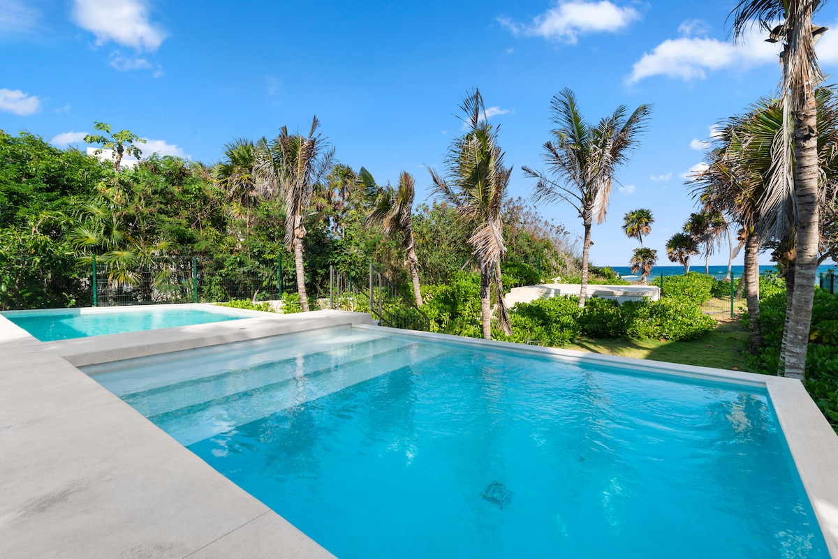 Two clear swimming pools are framed by a lush green landscape, with palm trees swaying gently in the breeze. The bright blue sky adds to the serene atmosphere, contrasting beautifully with the vibrant greenery surrounding the area.