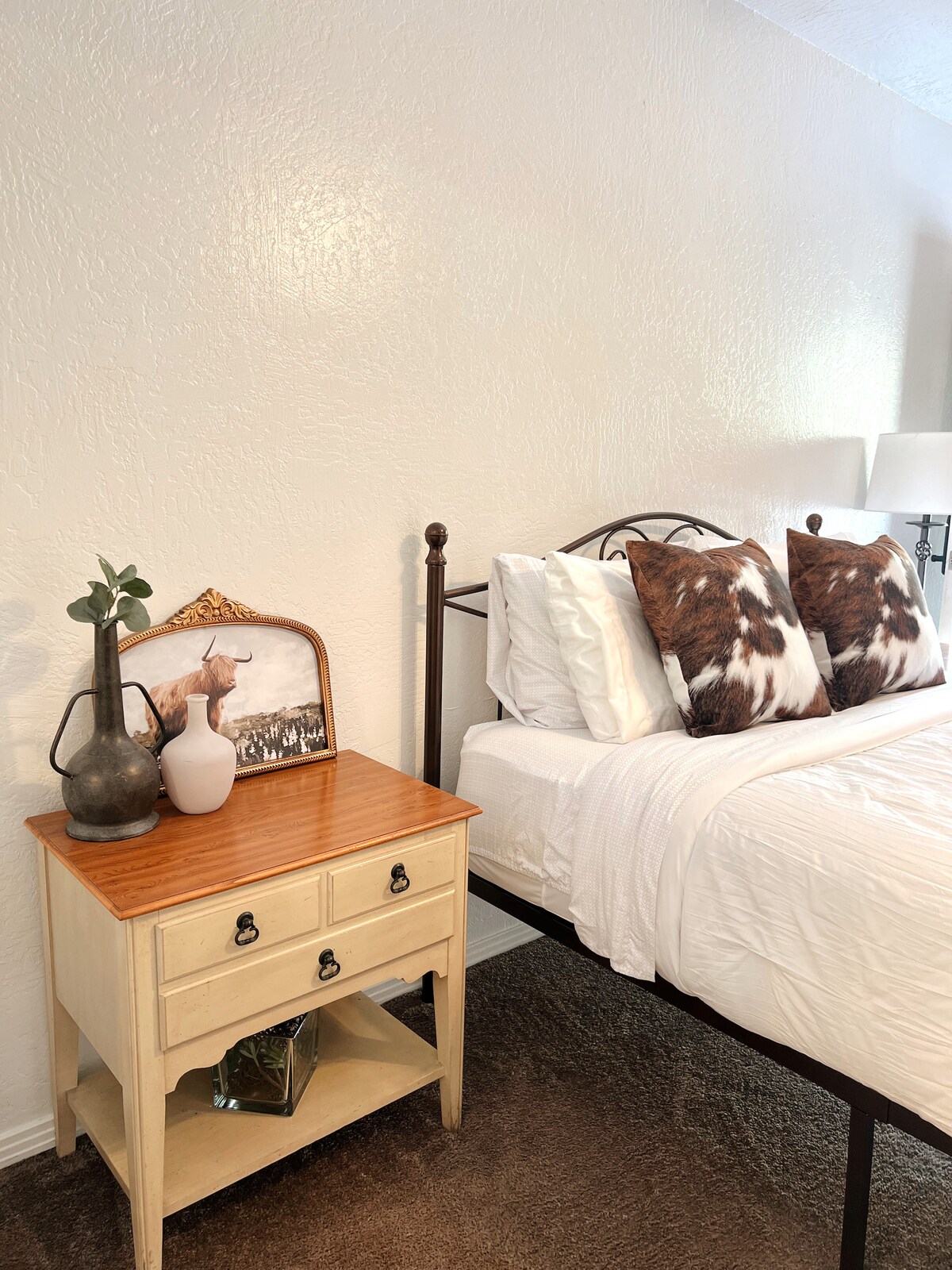 A cozy sleeping area features a sleek metal bed with two decorative pillows. A nightstand holds a vintage vase and a framed picture. Soft light is provided by two lamps on either side, complemented by warm wall tones and a textured carpet.