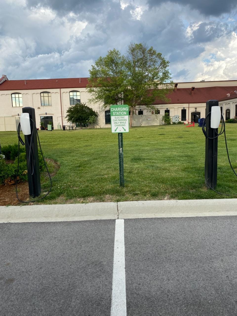 A designated charging station is displayed on a green sign, surrounded by a grassy area. Two charging units are positioned on either side of the sign, providing electric vehicle charging capabilities. The background features a building with a red roof and partially cloudy skies above.