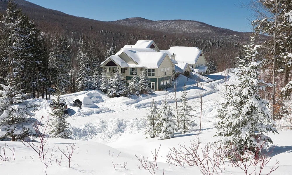 A spacious mountain retreat is nestled among snow-covered trees, surrounded by a winter landscape. The building features a combination of gabled roofs and large windows, blending harmoniously with the serene environment. Fresh snow blankets the ground, adding to the picturesque tranquility.