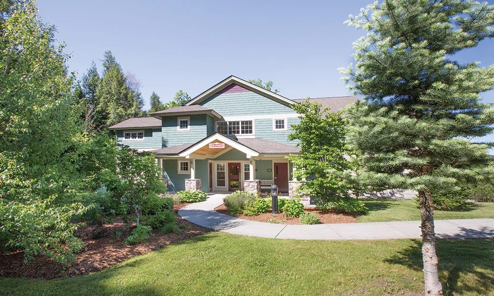 A spacious exterior view of a multi-story building surrounded by trees and well-maintained landscaping. A wide pathway leads to the entrance, bordered by lush greenery and shrubs, creating a welcoming atmosphere. The blue and green siding complements the natural surroundings.
