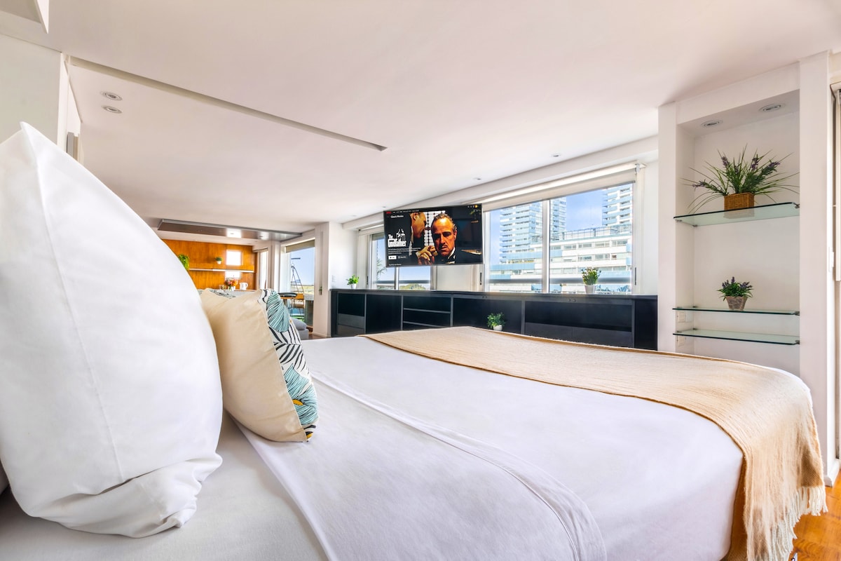 A spacious studio is shown, featuring a king-size bed with a light-colored comforter and large pillows. A 52-inch smart TV is mounted on the wall opposite the bed. Natural light floods the room through large windows lined with greenery.