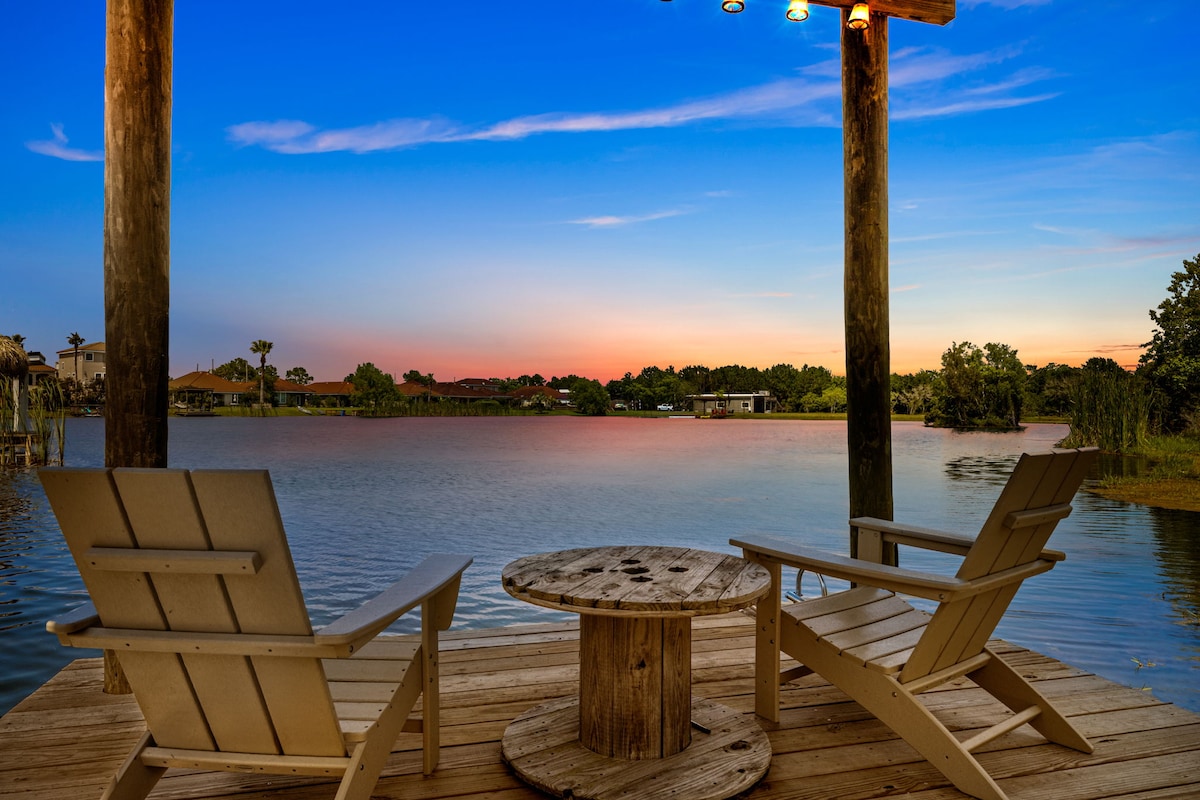 A wooden dock features two light-colored Adirondack chairs positioned beside a round table. The serene lake view is framed by trees and reflects the soft colors of the sunset sky, creating an inviting outdoor space for relaxation.