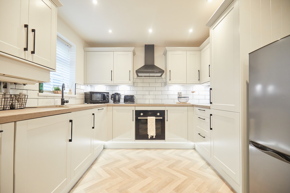 A modern kitchen space is featured, showcasing elegant cabinetry in a light finish. A built-in oven and stovetop are visible, complemented by a sleek, stainless steel refrigerator. The flooring consists of light wooden planks arranged in a herringbone pattern, enhancing the overall spaciousness.