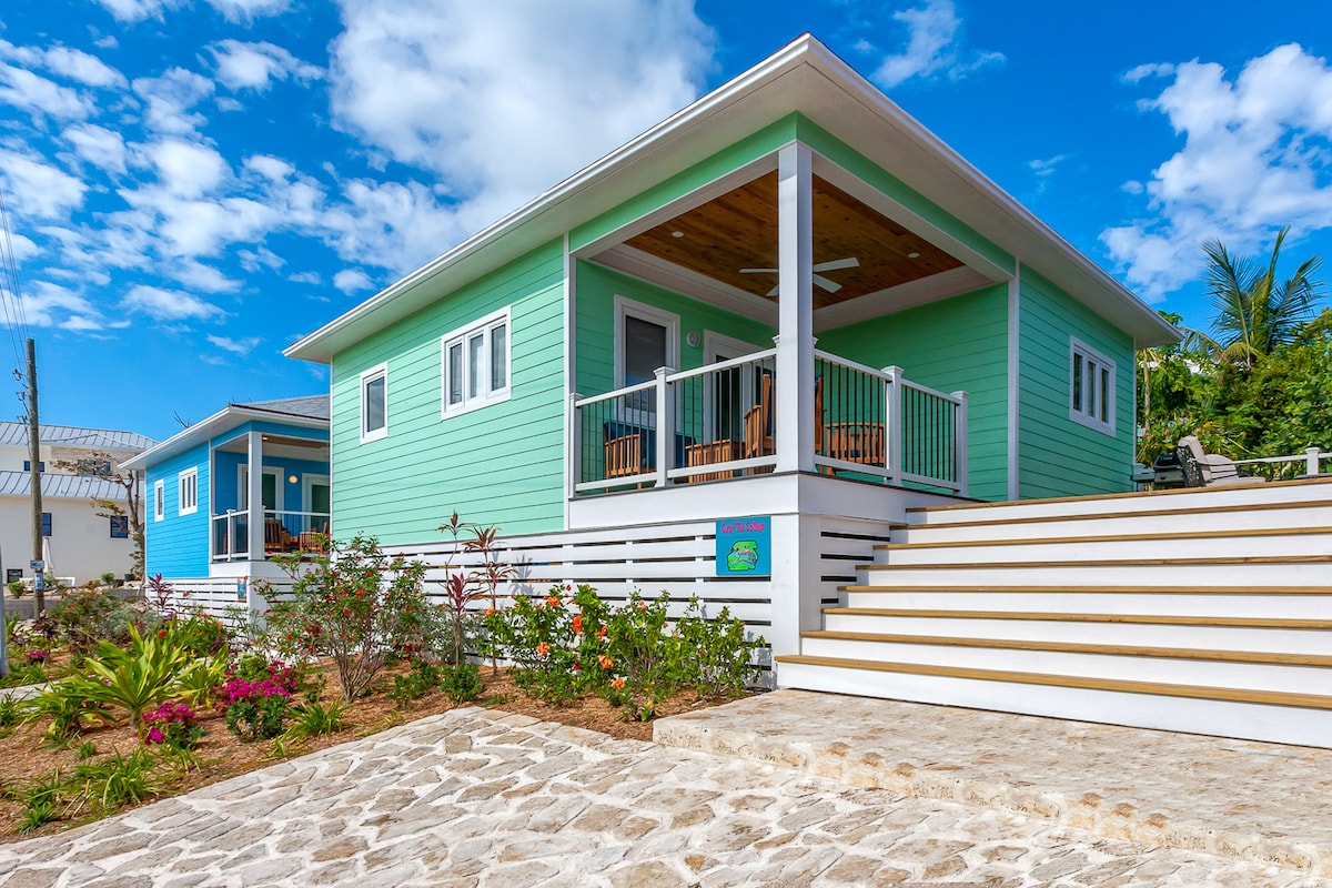 A modern cottage with a pastel green exterior is showcased, featuring a wooden porch furnished with rocking chairs. Steps lead to a landscaped area with colorful flowers and stone pathways, under a bright blue sky partially adorned with clouds.