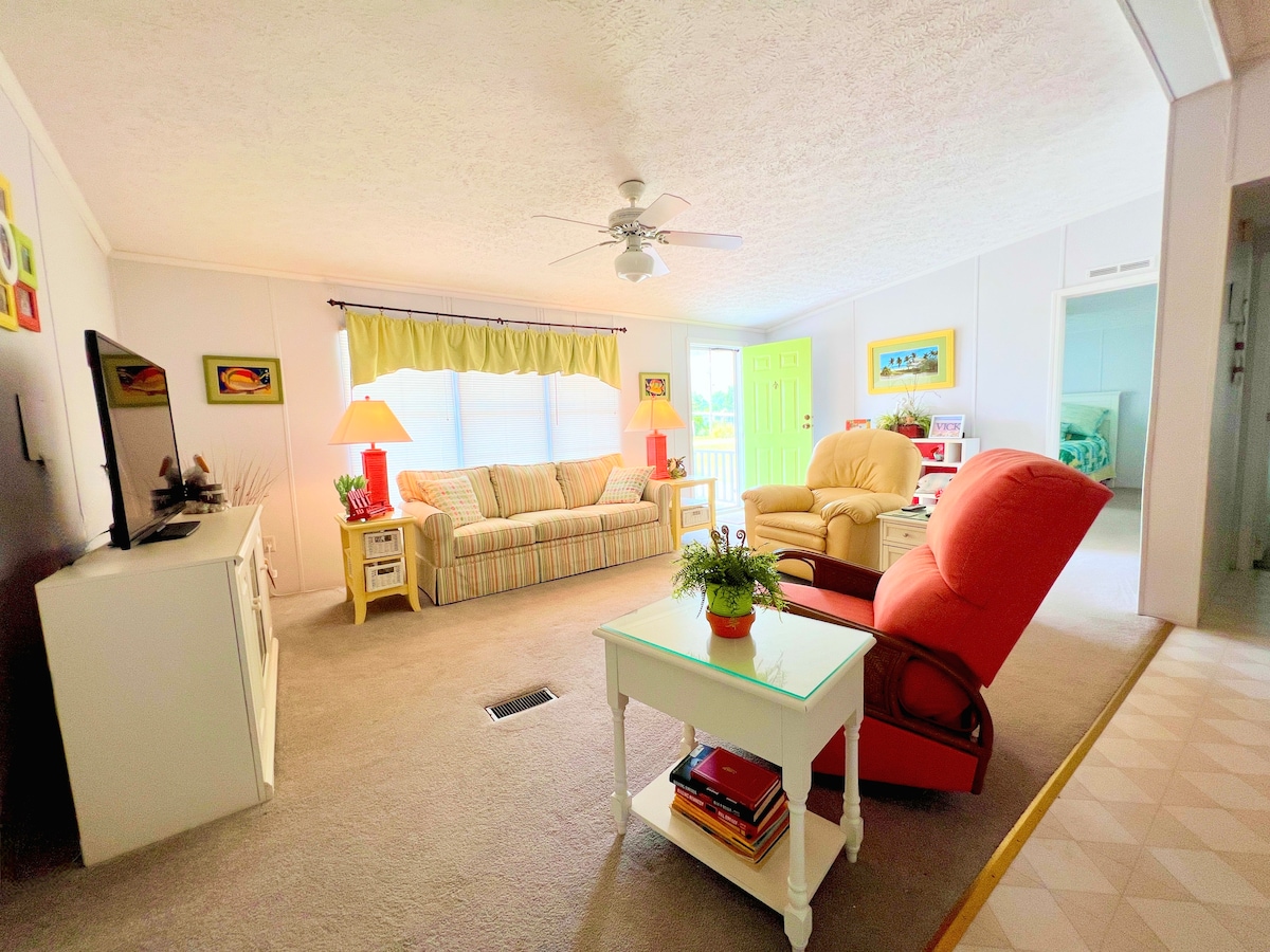 A bright living area features a combination of comfortable seating, including a striped sofa and a reclining chair. Natural light floods the space through large windows, while a ceiling fan provides a gentle breeze. A small coffee table sits in the center, adorned with a potted plant.
