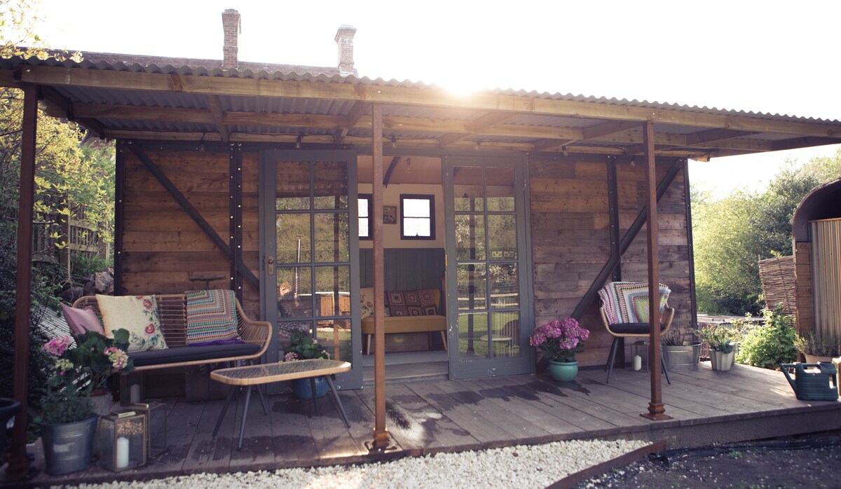 A charming wooden exterior is highlighted by a covered veranda, featuring seating areas adorned with colorful cushions. Sunlight filters softly through the open doors, creating a warm atmosphere. Planters of vibrant flowers add a touch of nature to the welcoming entrance.