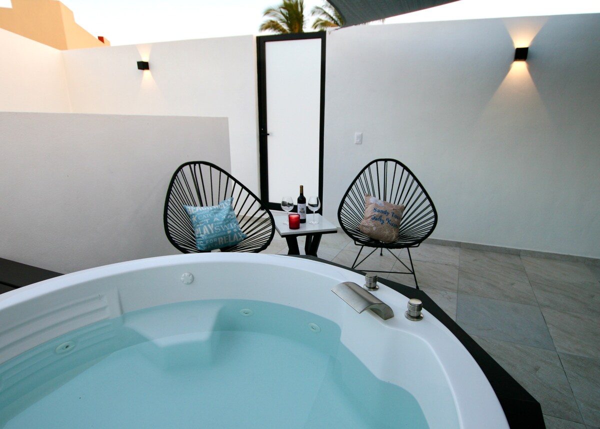 A private outdoor patio features a plunge pool with soothing blue water. Two black chairs with cushions are arranged around a small table, which holds a candle and two glasses. Soft lighting enhances the serene environment.