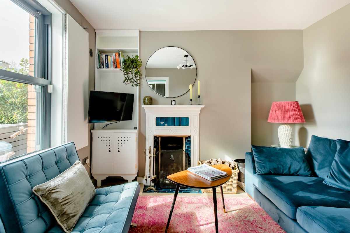 A comfortable living area is highlighted by a stylish blue sofa and a light blue loveseat. A round mirror hangs above a decorative fireplace, while a coffee table sits at the center. A pink rug adds warmth, and a lamp with a red shade provides soft lighting.
