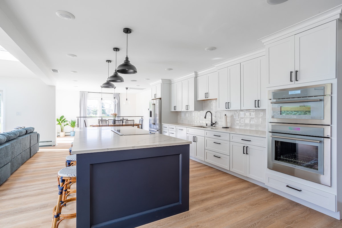 A spacious kitchen showcases a large island with seating for four and high-end appliances. The clean, white cabinetry contrasts with dark accents. Natural light floods the area, enhancing the modern, farmhouse style while providing a seamless connection to the dining area and living room.