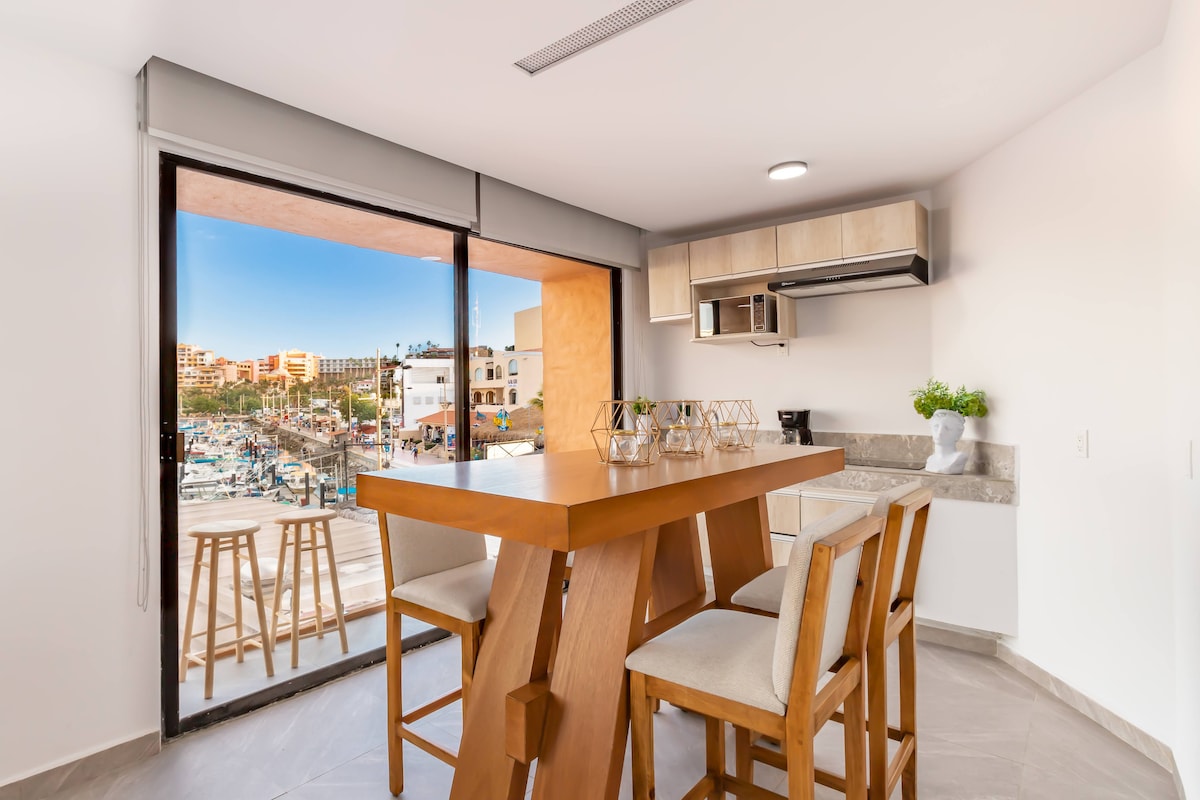 A modern dining area features a large wooden table with seating for four and stylish bar stools. Large windows allow natural light in, offering a view of the marina. The kitchen area includes sleek cabinetry and a countertop, enhancing the open layout.