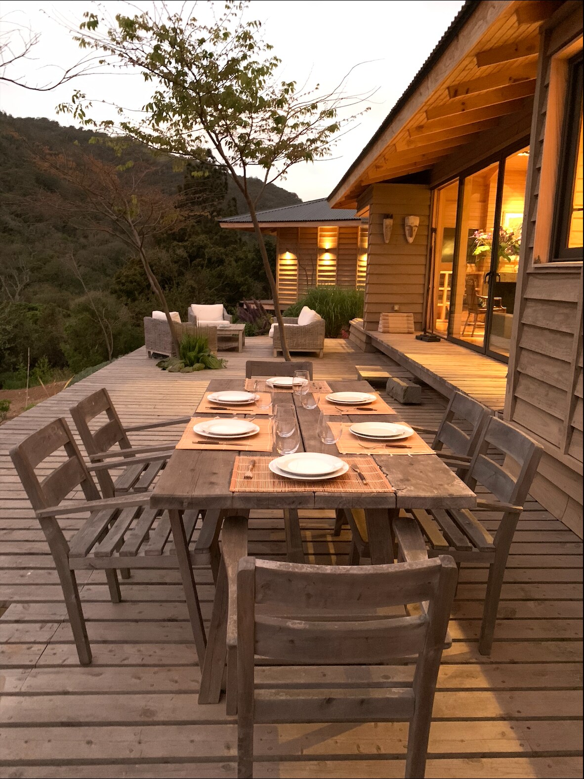 An outdoor dining area features a rustic wooden table set for six, surrounded by wooden chairs. Soft lighting highlights the natural surroundings, with a view of the hills beyond. The adjacent deck is lined with plants, creating a serene and inviting space.