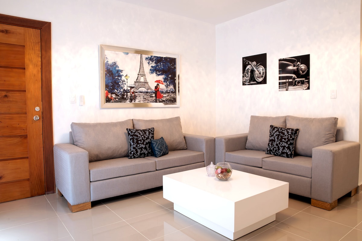A bright living area is presented, featuring two contemporary sofas upholstered in gray fabric. A sleek white coffee table sits in the center, adorned with a decorative bowl. Black and white framed artwork complements the warm wooden door, creating a balanced and inviting space.