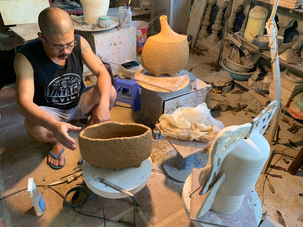 An artisan is seen working on ceramic pieces in a workshop. Various pots in different stages of completion are placed around the workspace, with tools and supplies evident. The environment suggests a creative and dynamic atmosphere, with natural materials and artistic objects present.