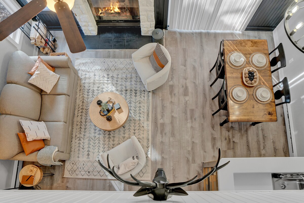 An overhead view of the open living area showcases a spacious couch and two club chairs arranged around a circular coffee table. A dining table with black chairs is set nearby. A wood-burning fireplace with a stone surround adds warmth to the room.