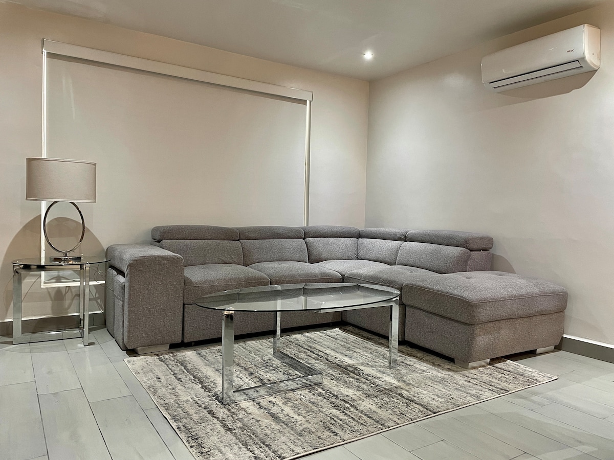 A modern living area features a spacious gray sectional sofa arranged around a glass coffee table. A textured area rug lies beneath, and ambient light is provided by a stylish lamp on an adjacent side table. Soft tones and clean lines create an inviting space.