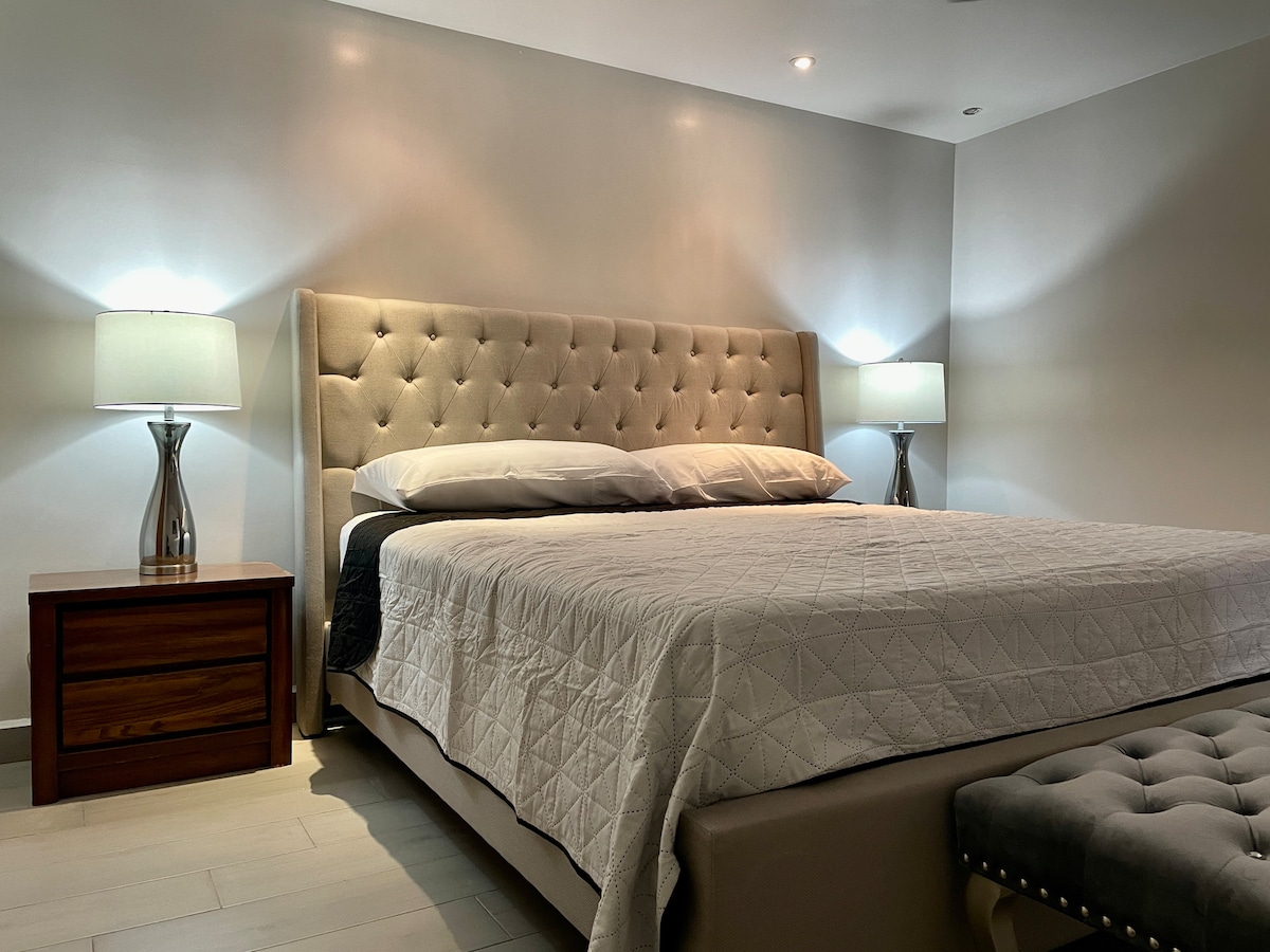 A spacious sleeping area features a tufted beige headboard with soft bedding. Two bedside lamps provide gentle illumination beside a wooden nightstand, creating a calm atmosphere. The room is minimally decorated, allowing for a serene and uncluttered environment.