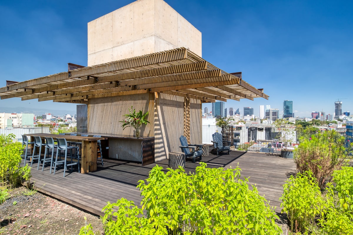 An expansive rooftop terrace features a wooden deck surrounded by greenery. A stylish bar area is complemented by several seating options, including modern stools and chairs. The backdrop showcases city views, revealing a mix of urban architecture under a clear blue sky.