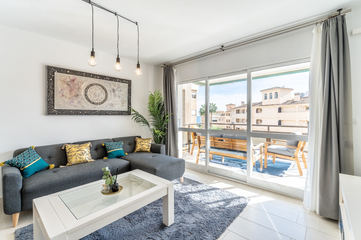 A modern living room features a gray sofa adorned with decorative pillows. Natural light fills the space through large glass doors that open to an outdoor terrace. A coffee table sits on a textured rug, and a small plant adds a touch of greenery.