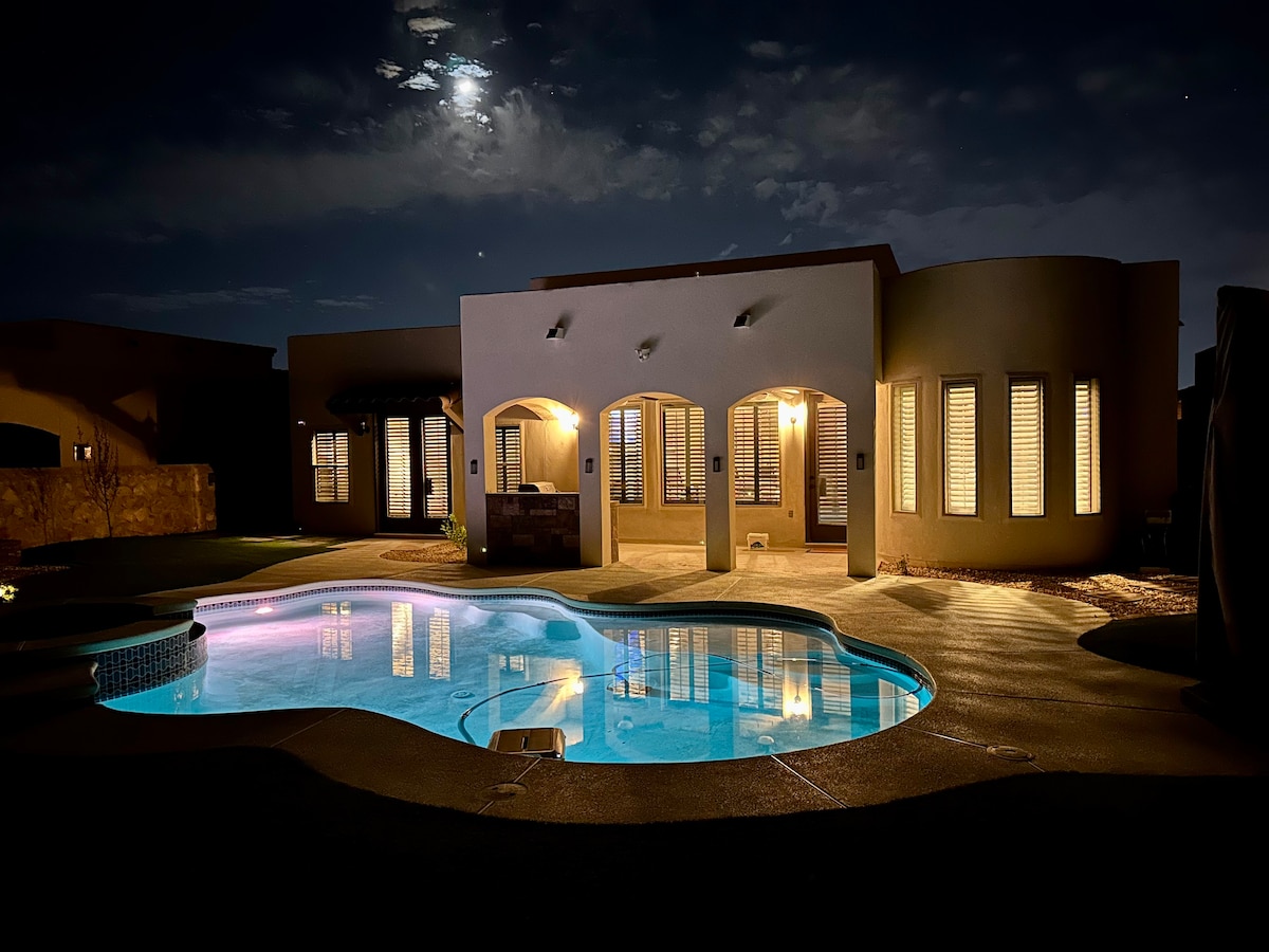 The exterior of the house is featured at night, showcasing a softly illuminated pool area. The building's warm light reflects on the water, while arched windows and decorative shutters create a serene ambiance under a partially clouded moonlit sky.