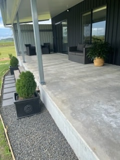 A covered veranda is shown featuring a spacious concrete floor. Two black outdoor chairs are placed near the building, accompanied by a potted plant. Decorative stones line the edge, providing a neat border alongside the greenery visible in the distance.