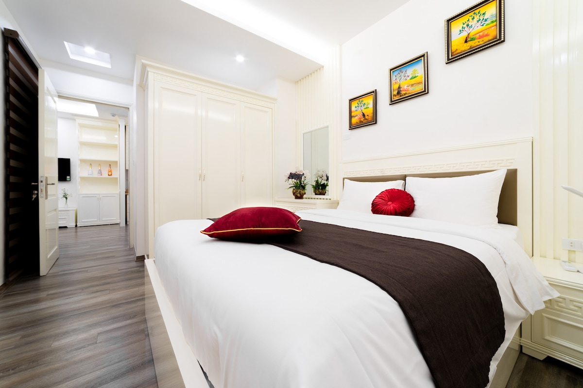 A well-appointed bedroom is presented with a spacious bed adorned with a dark blanket and a decorative red pillow. Bright artwork hangs above a crisp white headboard. Natural light enters through an adjacent doorway, while built-in wardrobes contribute to the room’s neat appearance.