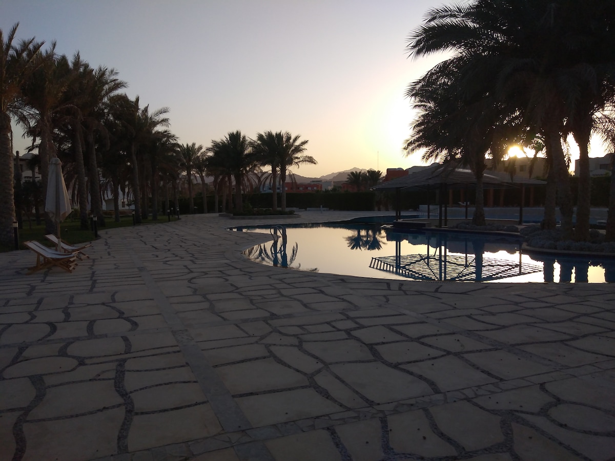 A serene pool area is surrounded by palm trees, reflecting the soft hues of the sunset. The stone-paved deck leads to a calm body of water, providing a tranquil setting with a gazebo structure visible in the background.