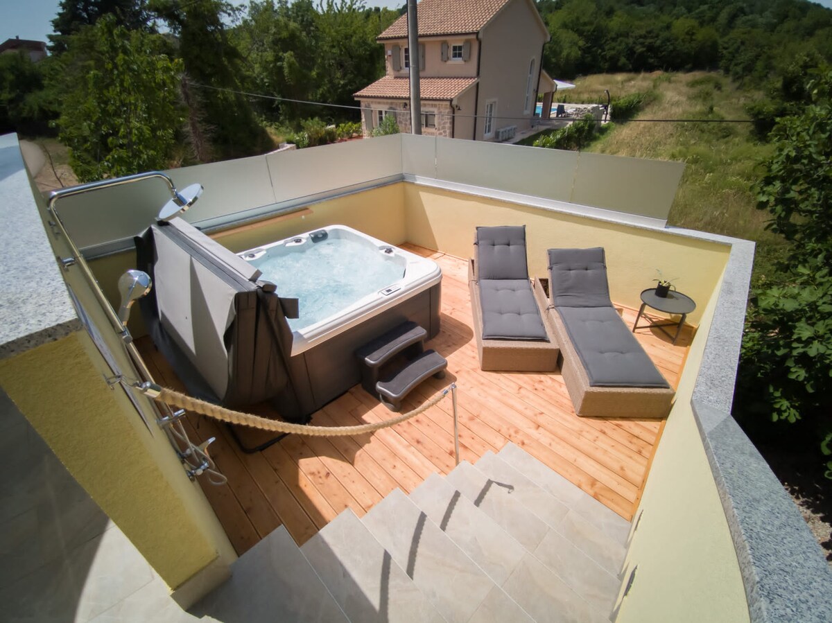 A private outdoor area features a hot tub surrounded by a wooden deck. Two lounge chairs provide relaxation space, accompanied by a small side table. The area is enclosed by low walls, offering a sense of seclusion, with greenery visible in the background.