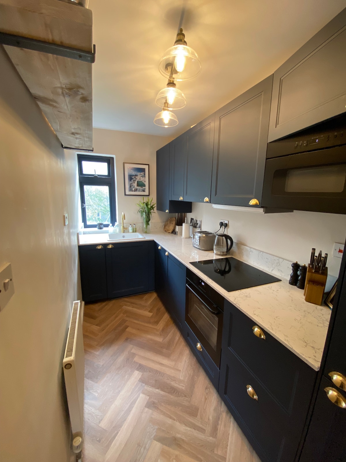 A modern kitchen is shown with navy blue cabinetry and marble countertop. Warm light is provided by hanging bulbs. An oven and microwave are integrated into the cabinetry, while a kettle and utensils are neatly arranged, emphasizing a functional yet stylish cooking space.