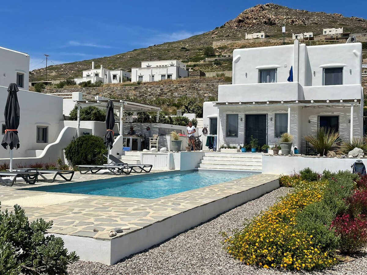 The exterior of a modern villa features a private pool bordered by sun loungers and umbrellas. Lush gardens with colorful flowers enhance the setting, while a hillside with additional white structures is visible in the background. Steps lead to the entrance of the villa.