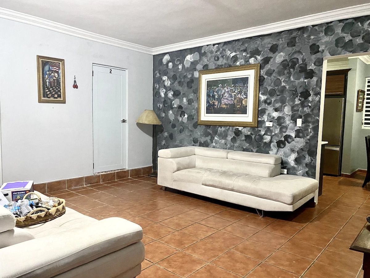 A spacious living area is presented, featuring two light-colored sofas arranged thoughtfully. The walls are adorned with abstract art, complemented by a framed piece. A lamp stands in one corner, and a door to the left leads to another space, while a textured gray wall adds character.