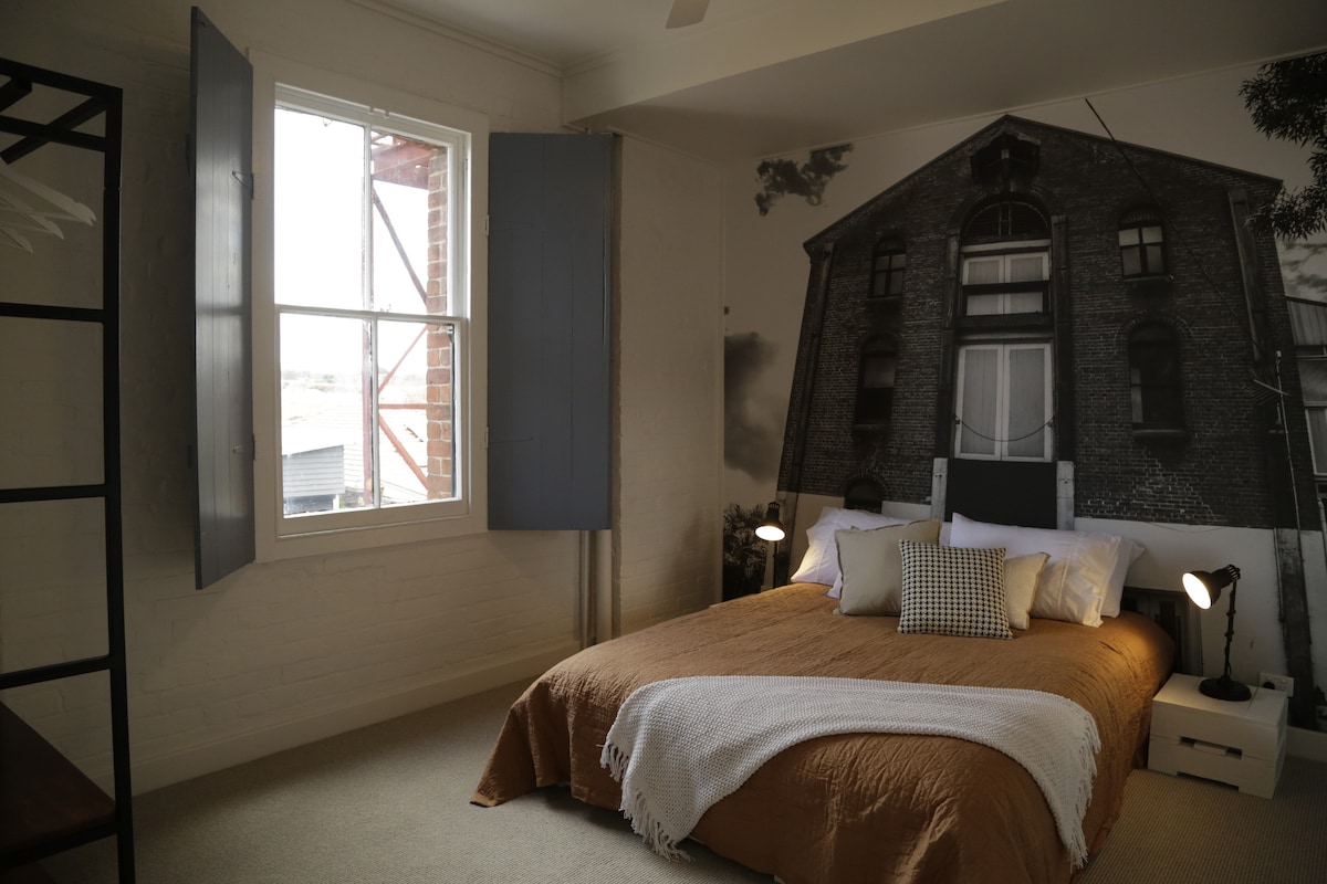 A spacious bedroom features a large bed with neutral bedding, accented by decorative pillows. An oversized wall mural showcases a historical building, while a window with wooden shutters allows natural light to enter the room. Two bedside lamps provide additional lighting.