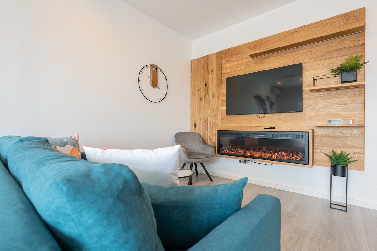 A modern living space features a cozy teal sofa, positioned toward a sleek wall-mounted television above an electric fireplace. Natural wood accents and a round wall clock contribute to a serene atmosphere. A minimalistic chair sits nearby, enhancing the inviting setting.