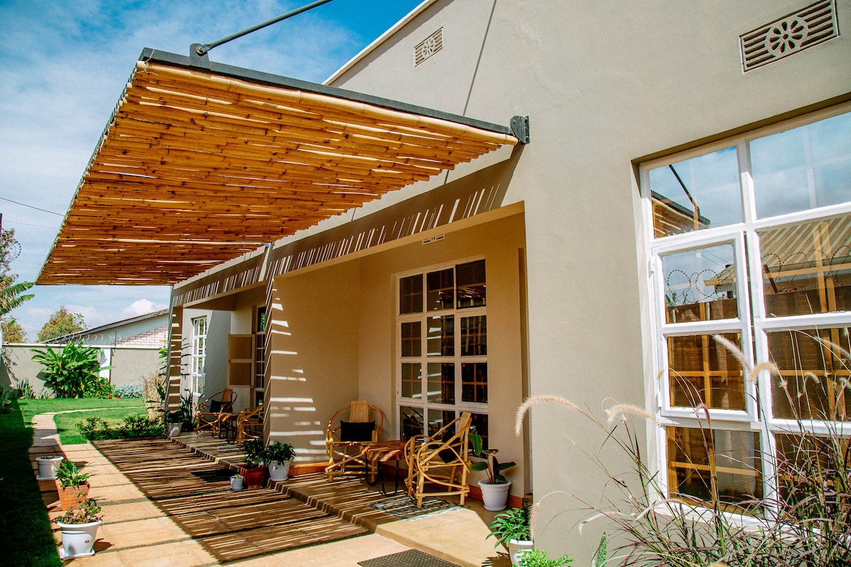 An outdoor area is showcased, featuring a wooden overhang that casts shadows on a spacious wooden deck. The deck is lined with potted plants and has several wooden chairs inviting relaxation. Large windows reflect natural light, complementing the fresh and inviting environment.