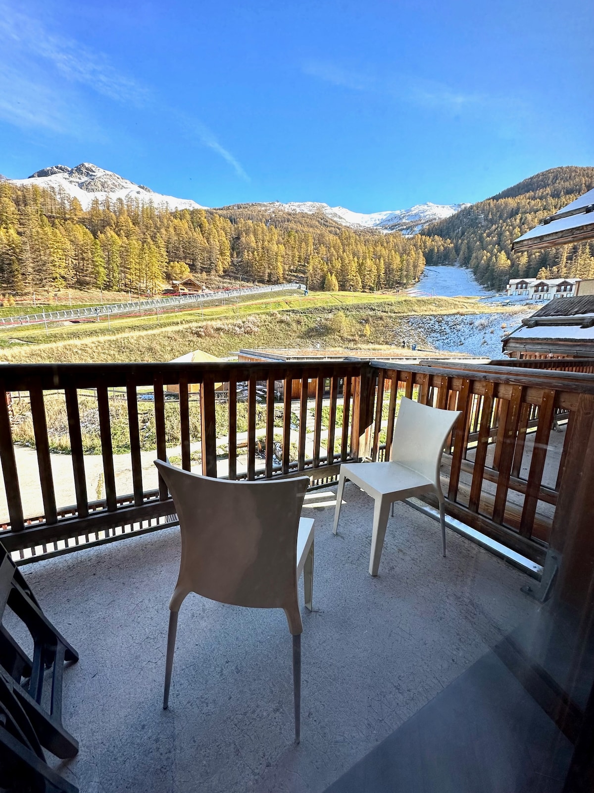 A balcony is featured with two white chairs overlooking a snow-covered landscape flanked by trees and mountains. The scene captures clear blue skies, creating a serene outdoor space for relaxation.