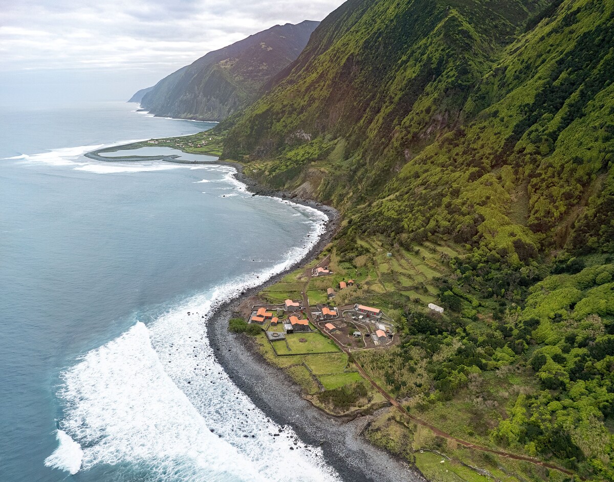 An aerial view captures the rugged coastline with lush green hills meeting the ocean. The village is situated along the shore, featuring a cluster of buildings with orange roofs amidst a landscape of greenery and rocky beach, creating a serene natural setting.
