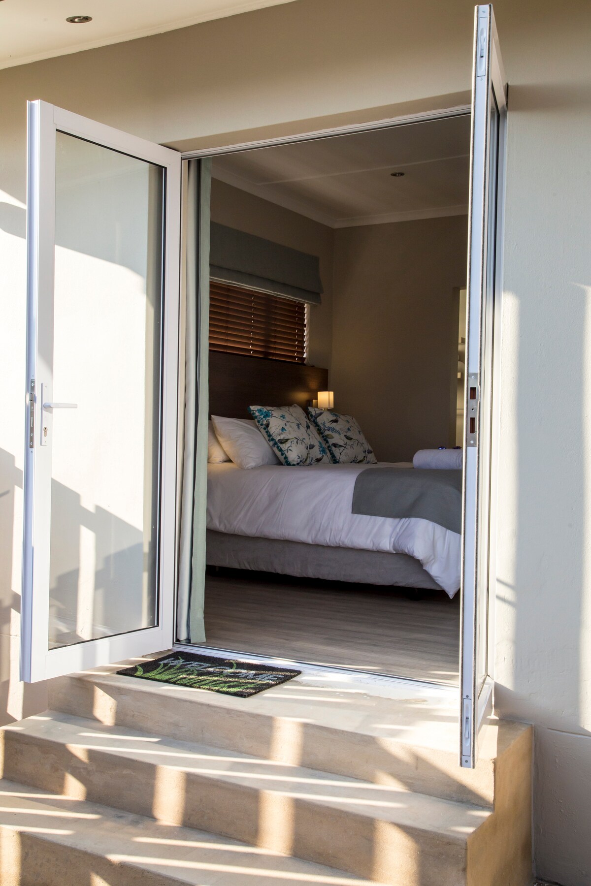 A private entrance is showcased, leading to a cozy suite. Light enters through the open doors, revealing a well-appointed sleeping area with a spacious bed adorned with subtle decor. Steps guide the way up to the entrance, with a welcome mat adding a personal touch.