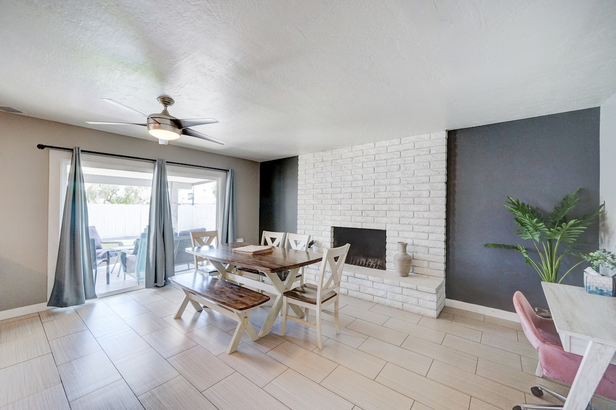 A spacious dining area features a large wooden table surrounded by white chairs. A stone fireplace stands against a dark wall, while large windows with flowing curtains allow natural light to illuminate the room. A green plant adds a touch of nature to the space.