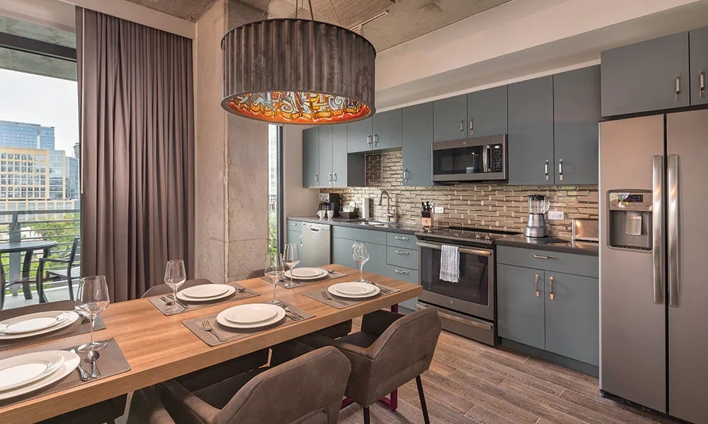 A modern kitchen is featured, with sleek gray cabinetry and a stylish island with seating. Stainless steel appliances, including a refrigerator and microwave, are visible. Natural light enters through large windows, illuminating the adjacent dining area with a wooden table set for six.