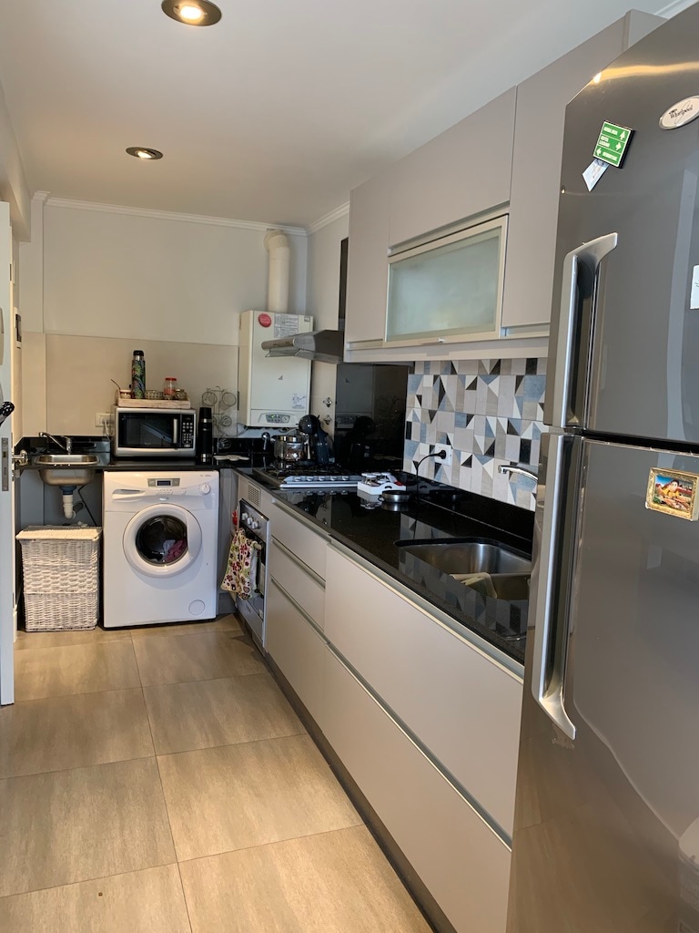 A modern kitchen is presented with sleek cabinets and a dark countertop. Essential appliances are visible, including a refrigerator and microwave. A washing machine is positioned beside the sink, while patterned tiles add interest to the space. Ample light is provided by overhead fixtures.