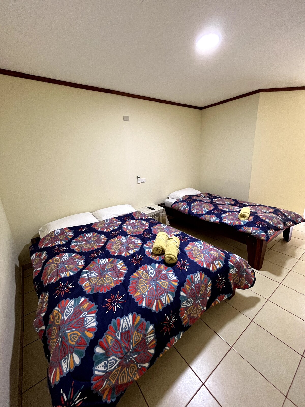 The sleeping area features two spacious beds, each covered with colorful bedspreads. Neutral-toned walls provide a calming backdrop. A small nightstand sits between the beds, with a few neatly rolled towels placed atop it.